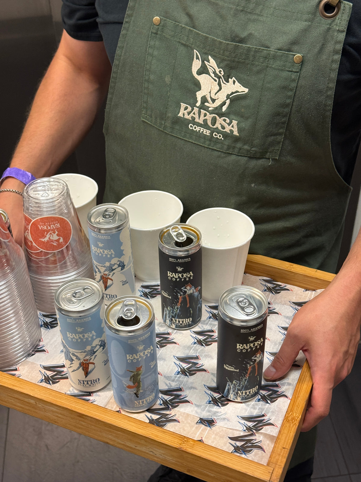 A barista in a Raposa apron carrying a tray of Raposa Nitro cans and cups